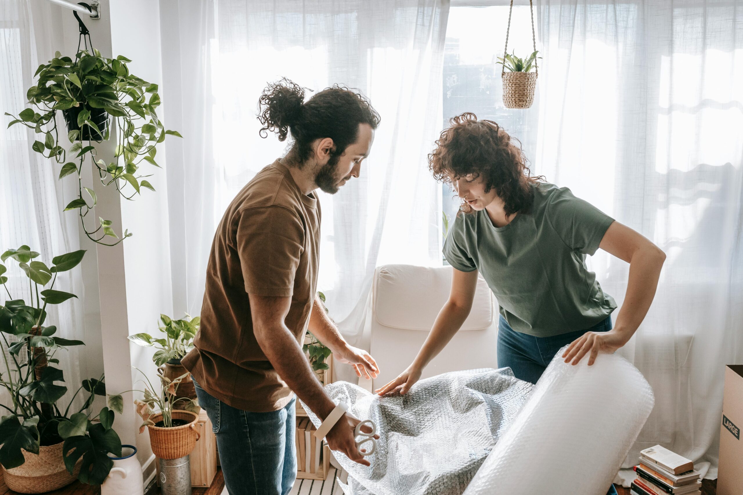 A couple wraps furniture in a plant-filled living room, preparing to move.